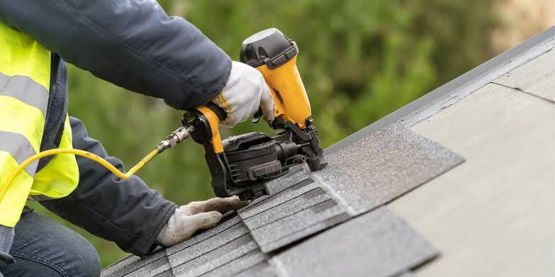 Roofer in a high-visibility vest nailing a shingle course with a pneumatic nailer